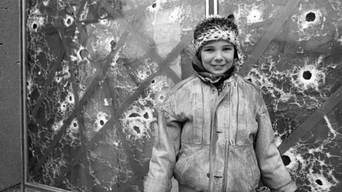 Bambina che sorride fotografata davanti a una vetrata perforata da pallottole