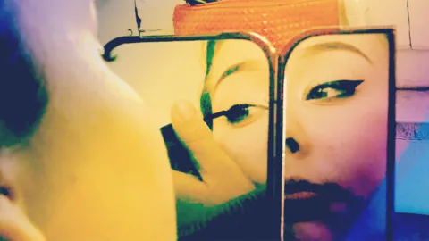 Ragazza con capelli scuri si trucca gli occhi riflessa in uno specchio posato su un tavolo